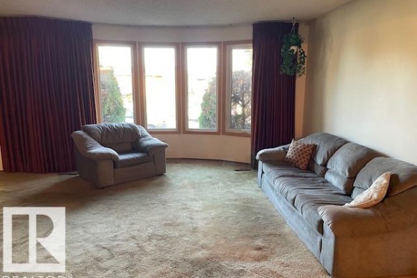 Living room with a grey sofa, grey armchair, beige carpet, and large bay window. - 8305 100 Av, Fort Saskatchewan