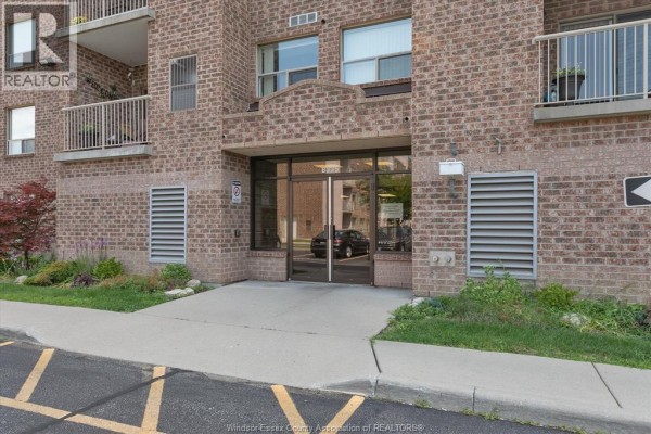 Entrance to the apartment building with a glass door, brick facade, and landscaping. - 8335 Wyandotte Street East Unit# 108, Windsor