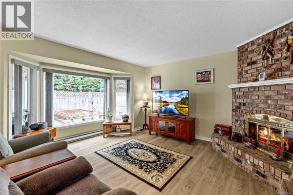 Living room with a bay window, wood-burning brick fireplace, television, and wood-look laminate flooring. - 834 Redonda Pl, Qualicum Beach