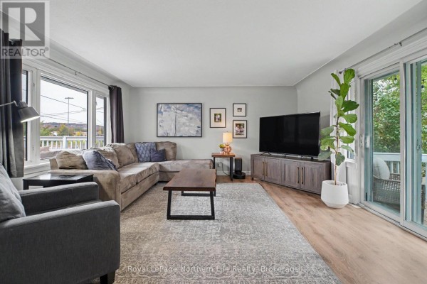 Living room with a sectional sofa, coffee table, TV stand, large windows, and a sliding door. - 835 Lorne Avenue, North Bay