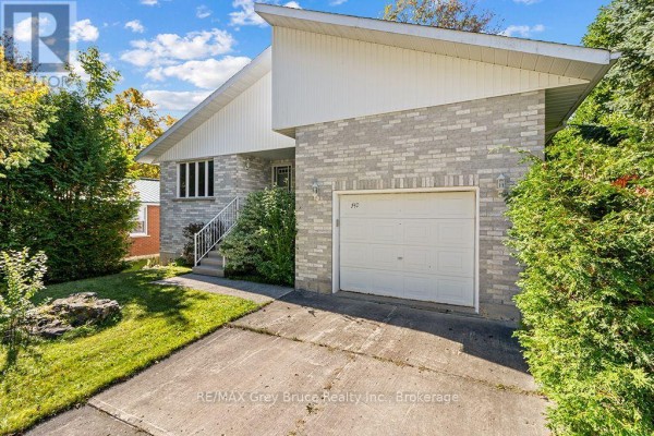 Side view of a grey brick house with a white garage door and mature trees. - 840 Mcnaughton Street, South Bruce Peninsula