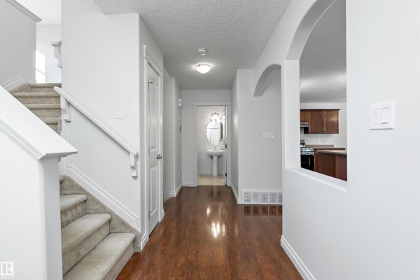 Interior view of a home hallway with hardwood floors, a carpeted staircase, and an archway to the kitchen. - 8419 2 Av Sw, Edmonton