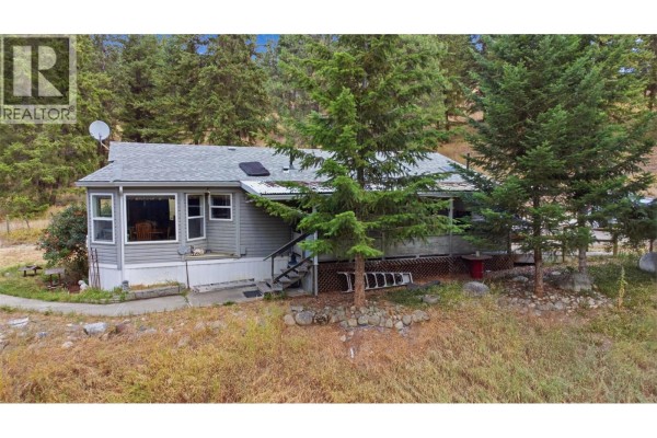 Front of the mobile home with a covered deck, steps, and mature trees in the yard. - 85 Candide Drive, Lumby