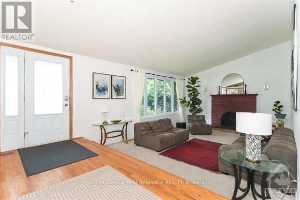 Living room with a brick fireplace, large window, plush seating, and hardwood floors. - 85 Crownhill Street, Ottawa
