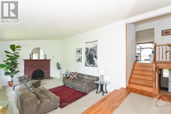 Living room with a brick fireplace, wooden staircase, and plush seating on carpeted floors. - 85 Crownhill Street, Ottawa