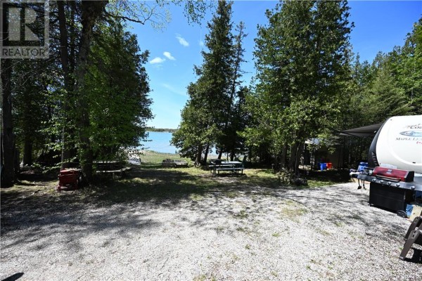 Gravel campsite area with trees, picnic tables, a grill, and a trailer, facing the lake. - 85 Leason Bay Trail, Assiginack