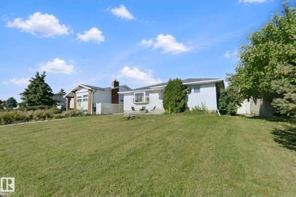 Side view of a white bungalow with a sloped lawn and mature trees under a cloudy sky. - 8508 149a Av Nw, Edmonton
