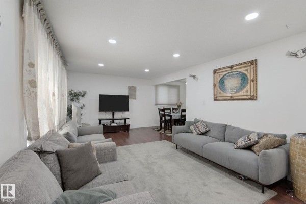 Living room with grey sectional sofa, television, dining table, hardwood floors, and large windows with sheer curtains. - 8508 149a Av Nw, Edmonton