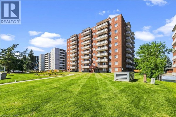 Red brick apartment building with balconies, green lawn, and trees under a blue sky. - 851 Queenston Road Unit# 202, Hamilton