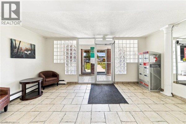 Apartment building lobby with beige tiled floor, glass block windows, seating, and mailboxes. - 851 Queenston Road Unit# 202, Hamilton