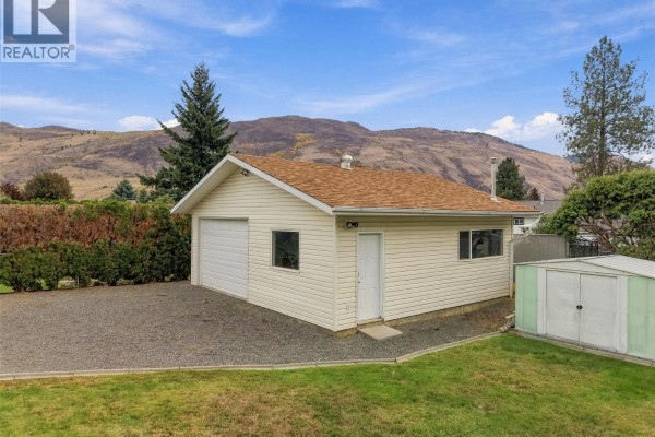 Exterior of a detached beige garage with a white roll-up door and two windows, set on a gravel pad. - 851 Sumac Place, Kamloops