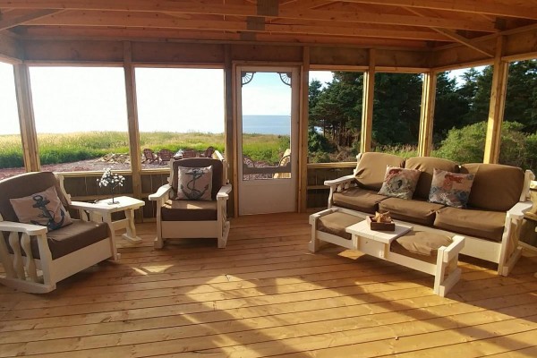 Interior of a screened-in porch with wooden furniture, pillows, and ocean view. - 8519 Route 14, Campbellton