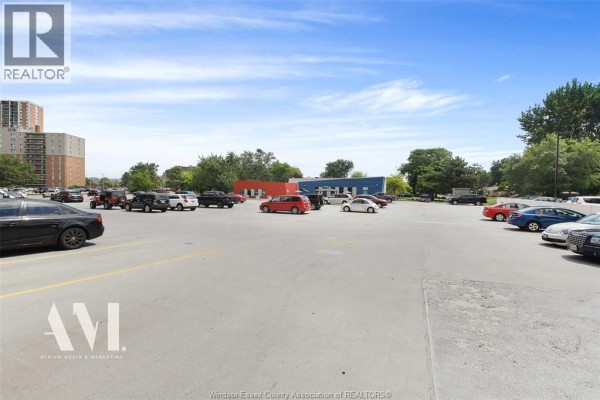 A view of a large, paved parking lot with cars and trees under a blue sky. - 8591 Riverside Drive East Unit# 308, Windsor