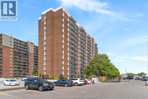 Exterior of a brick apartment building with a parking lot in front and trees to the side. - 8591 Riverside Drive East Unit# 702, Windsor