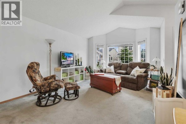 Living room with a vaulted ceiling, beige carpet, a sectional sofa, and bay windows overlooking houses. - 86 Coventry Lane Ne, Calgary