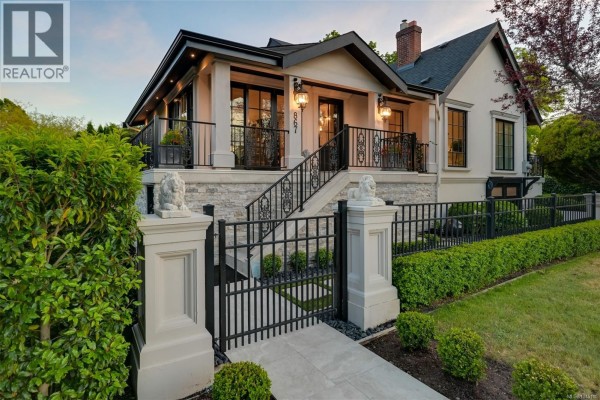 A wide view of the beige house's facade showcasing the entrance, stone base, black fence, landscaped hedges, and a paved pathway. - 867 Victoria Ave, Oak Bay