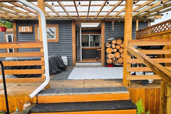 Outdoor seating area with a covered patio, firewood stack, and entrance to the mobile home. - 87 5250 Beaver Harbour Rd, Port Hardy