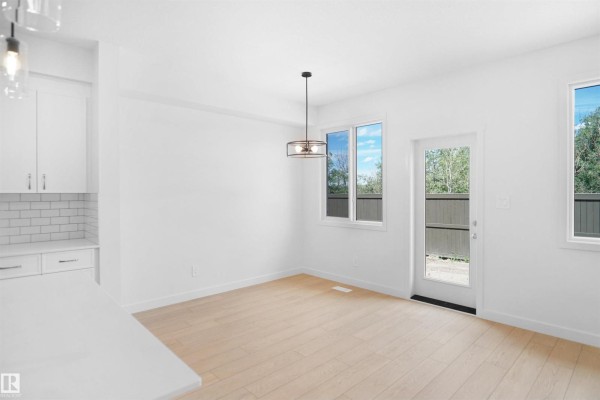 Dining area with white walls, light wood floors, a pendant light, and windows overlooking the backyard. - 8707 183 Av Nw, Edmonton