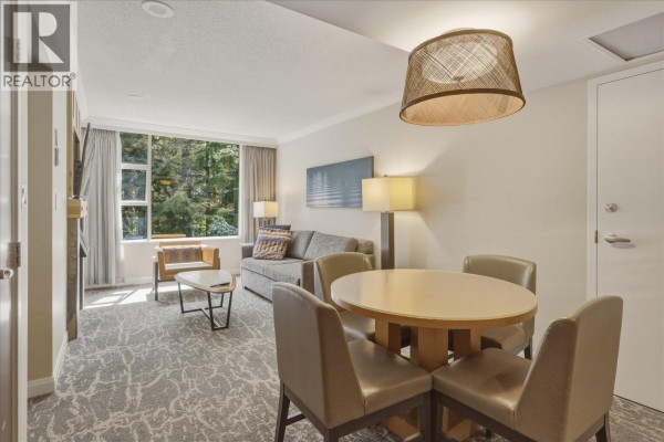 Living area with sofa, coffee table, and dining table with chairs, featuring a large window overlooking trees. - 871/872 4090 Whistler Way, Whistler