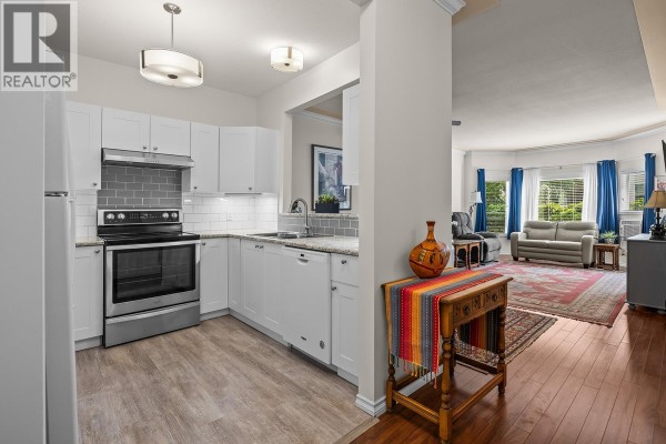 White kitchen with stainless steel appliances, grey tiled backsplash, and view into living room. - 877 Klo Road Unit# 112, Kelowna