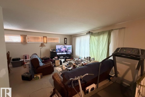 Living room with plush sofas, a treadmill, entertainment center, and large windows with blinds. - 8816 137 Av Nw, Edmonton