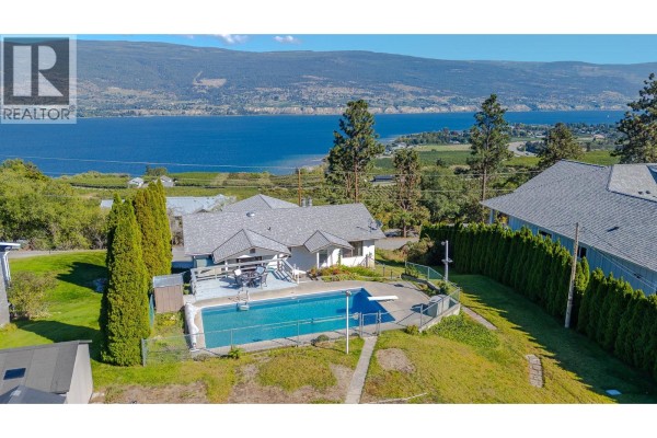 Rear view of a house with a swimming pool and deck, set against a scenic lake and vineyard landscape. - 8820 Happy Valley Road, Summerland