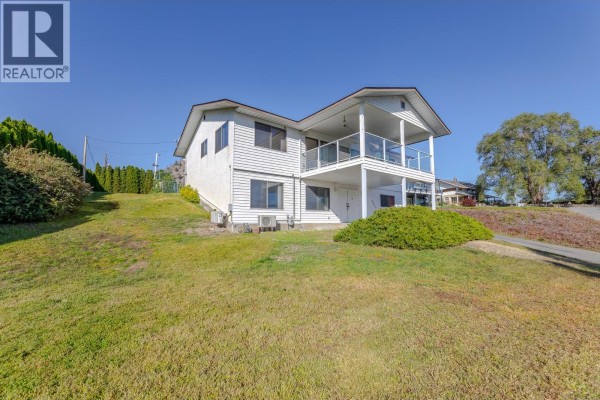 Side view of a white two-story house on a sloped lawn with mature trees and shrubs. - 8820 Happy Valley Road, Summerland