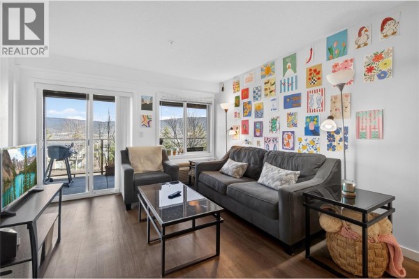 Living room with gray sofa, artwork on walls, and large windows overlooking the view - 883 Academy Way Unit# 116, Kelowna