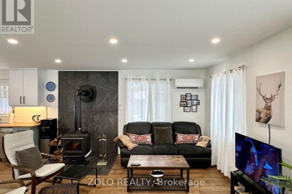 Living room facing the kitchen with white cabinets, black sofa, and wood-burning stove. - 886 Monck Road, Kawartha Lakes