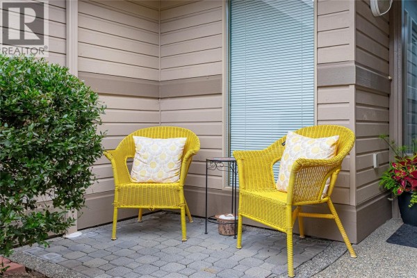 Two yellow wicker chairs with pillows sit on a paved patio area beside the townhouse entrance. - 888 Lakes Blvd, Parksville