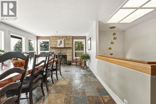 View of the dining room from the staircase landing, showing the table, fireplace, and windows. - 8905 Russet Drive, Vernon