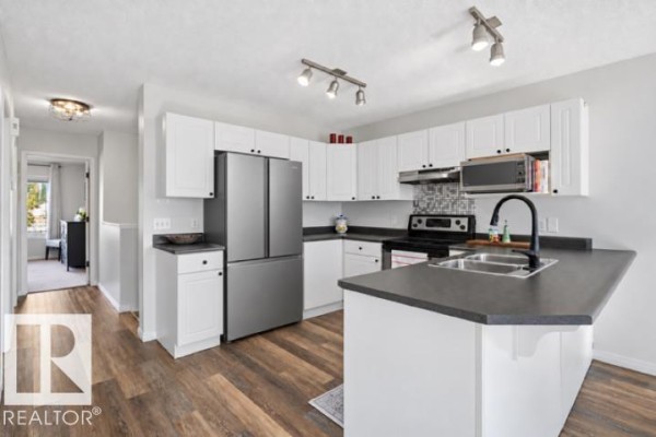 L-shaped kitchen with white cabinetry, stainless steel appliances, dark countertops, and a view into a hallway. - 8911 173 Av Nw, Edmonton