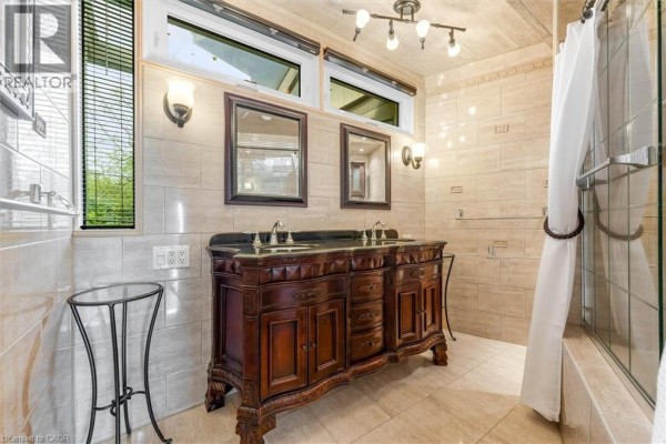 A large wooden vanity with a double sink and mirrors is featured in this tiled bathroom. - 899 Niagara Parkway, Fort Erie