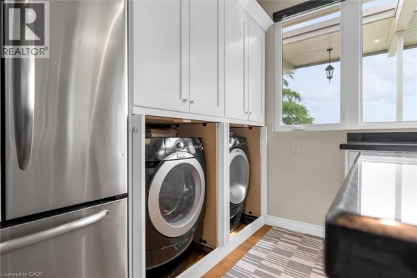 Laundry area with a stainless steel refrigerator, white cabinets, and stacked washer and dryer units. - 899 Niagara Parkway, Fort Erie