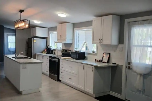 Wide view of a white kitchen with stainless steel appliances, island, and pendant light. - 9 Central Ave, Elliot Lake