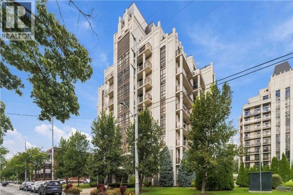 Side view of a tall, light-colored apartment building with balconies, surrounded by green trees. - 90 Charlton Avenue W Unit# 203, Hamilton