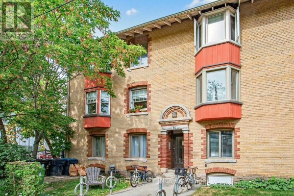 Front exterior of a yellow brick apartment building with bay windows, showing entrance and bicycles. - 90 Oxford Street, Toronto