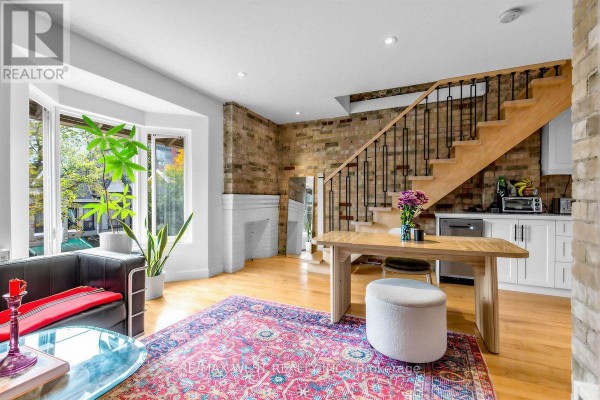 Living area with hardwood floors, exposed brick wall, bay window, dining table, and staircase. - 90 Oxford Street, Toronto