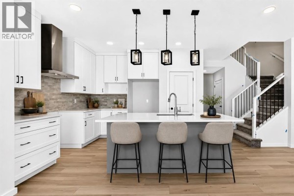 A modern white kitchen with a large island, pendant lights, stainless steel appliances, and hardwood floors. - 90 Raven Point Se, Calgary