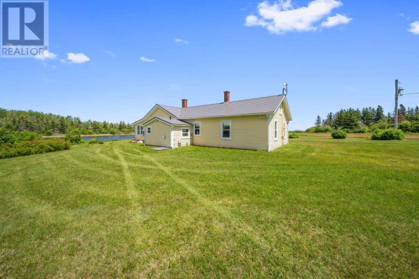 Side view of a yellow house surrounded by a grassy field and trees with water beyond. - 90 Tuplin Rd, Ellerslie Bideford