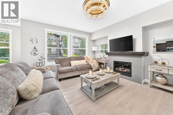 Living room with grey couches, a gas fireplace, large windows, and light wood flooring. - 901, 110 Coopers Common Sw, Airdrie