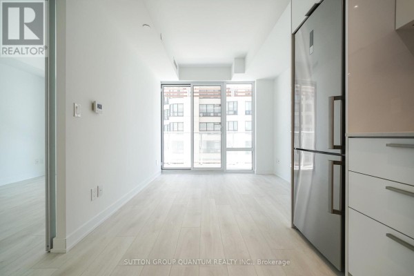 Empty apartment living area with light wood floors and large windows. Refrigerator visible on right. - 902 - 18 Maitland Terrace, Toronto