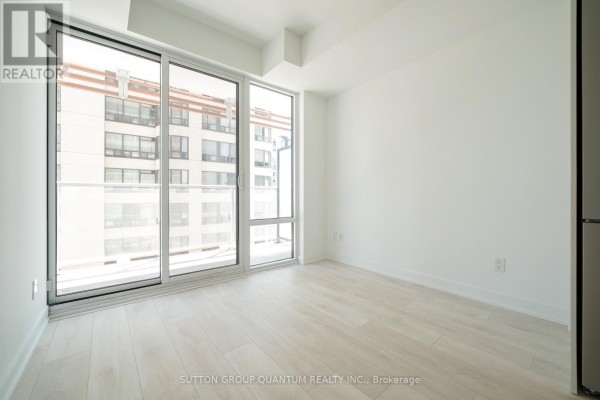 Empty apartment room with light wood floors and large windows offering a view of adjacent buildings. - 902 - 18 Maitland Terrace, Toronto