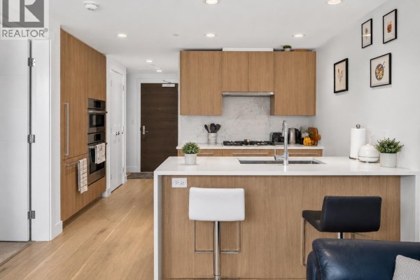 Close-up of a kitchen with gas stove, white backsplash, sink, and light wood cabinets. - 902 1210 E 27th Street, North Vancouver