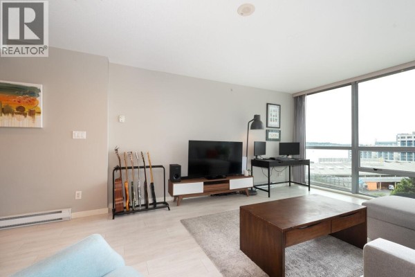Living room with a TV, desk, sectional sofa, coffee table, and a large window overlooking the city. - 902 14 Begbie Street, New Westminster