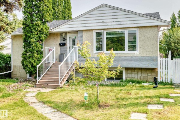 A suburban house with light beige textured siding, stone foundation, and a white picket fence. - 9031 150 St Nw, Edmonton