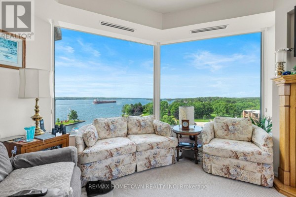 Living room with floral sofas and large windows overlooking a wide blue river and green trees. - 904 - 15 St Andrew Street, Brockville