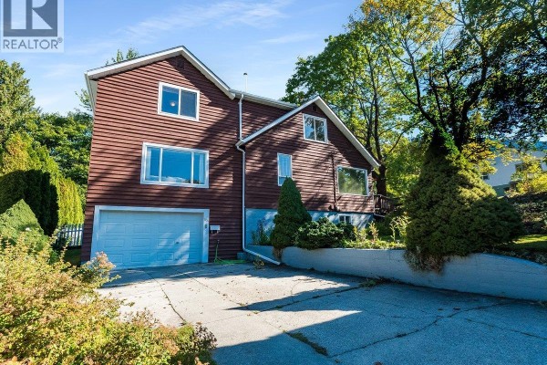 Driveway view of a house with brown siding, a garage, and surrounding greenery. - 904 Fifth Street, Nelson