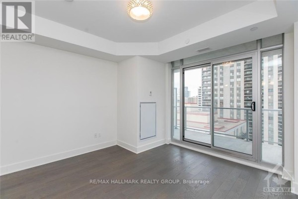 Spacious room with dark wood floors, white walls, and a sliding glass door to a balcony. - 905 - 255 Bay Street, Ottawa