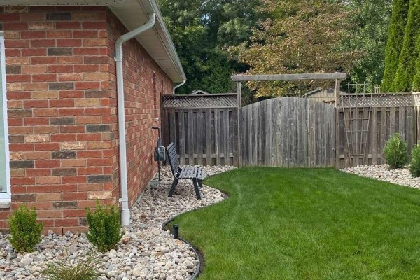 Backyard view with a wooden fence, green grass, a black bench, and a pathway through rocks. - 9053 Indian Creek Line E, Chatham Kent